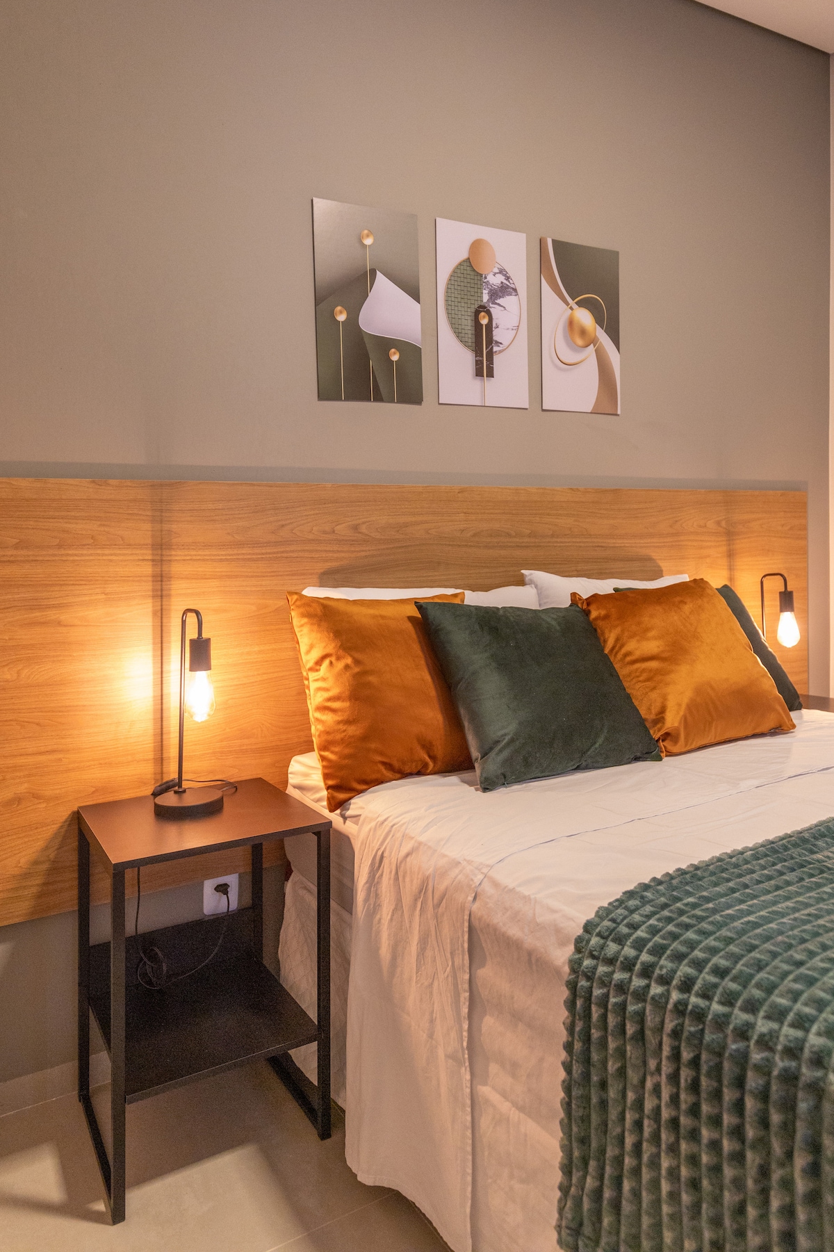 The sleeping area features a spacious bed with a light-colored duvet, accented by rich green and orange pillows. Minimalist artwork hangs above the headboard, while bedside lamps provide soft lighting, enhancing the modern design of the room.