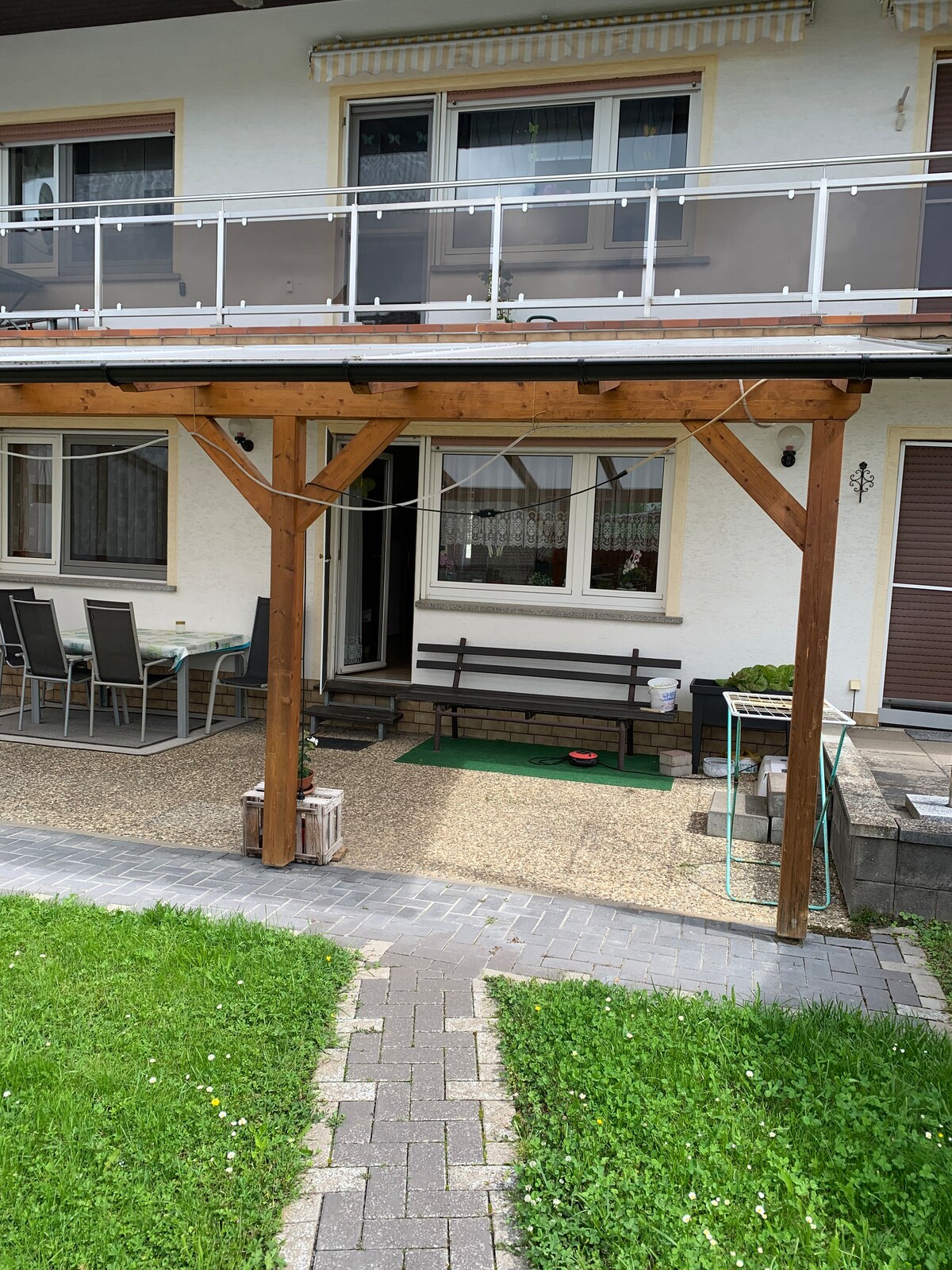 A covered terrace area is visible, featuring a wooden structure with a gravel floor. Several chairs surround a table, and a bench is positioned nearby. The entrance to the apartment can be seen in the background, with green grass bordering the stone pathway.