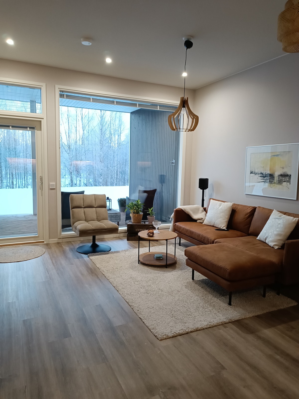 A modern living area features a brown sectional sofa and a glass coffee table on a soft area rug. A comfortable chair is positioned nearby. Large glass doors open to a snowy view outside, creating a bright and inviting space. Decor includes indoor plants and artwork on the wall.