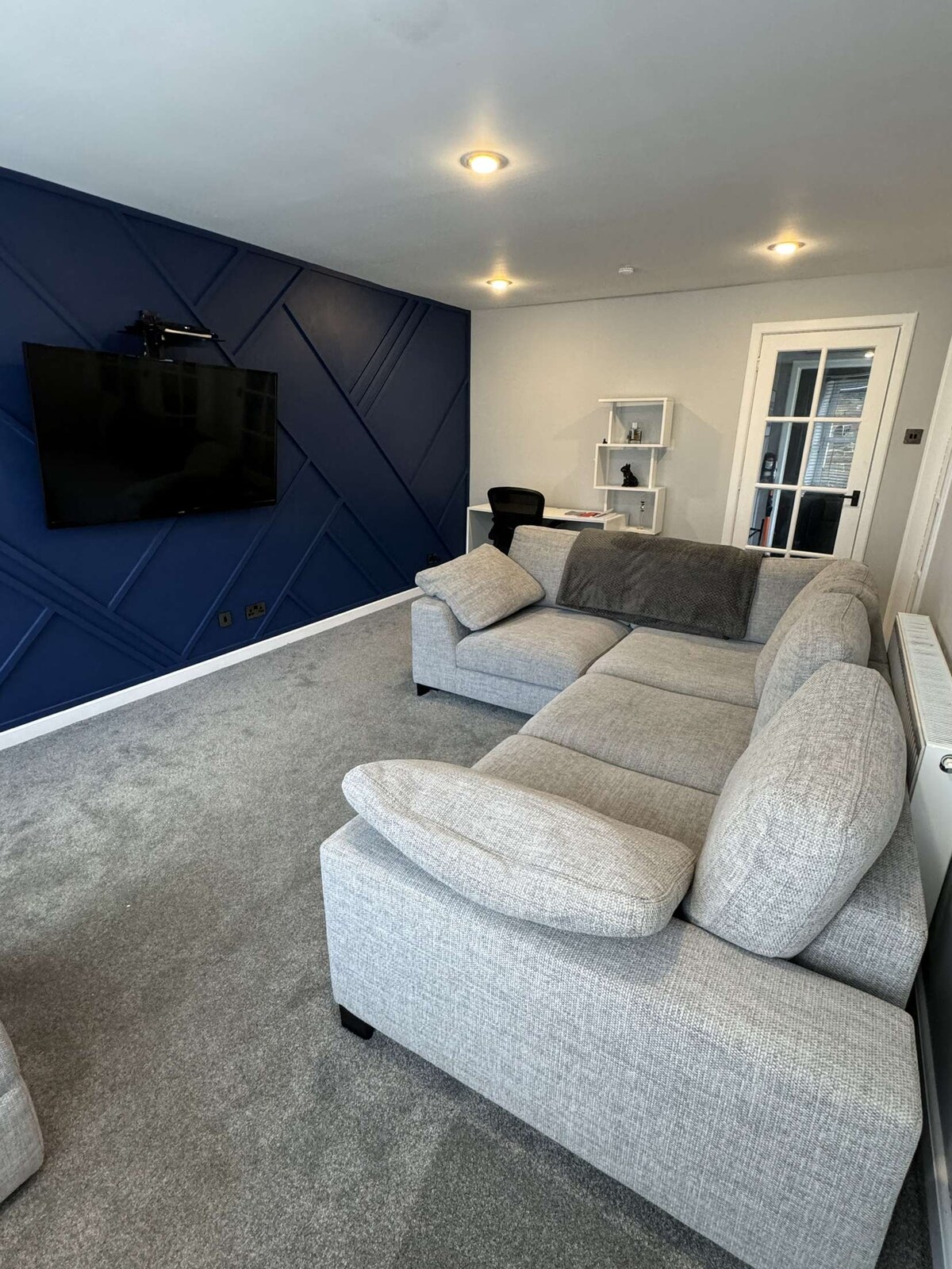 A living area is shown featuring a light gray sectional sofa with oversized cushions. A dark blue accent wall adds depth to the space. A flat-screen TV is mounted above a simple media console. Natural light is present through a glass door leading to an adjoining room.