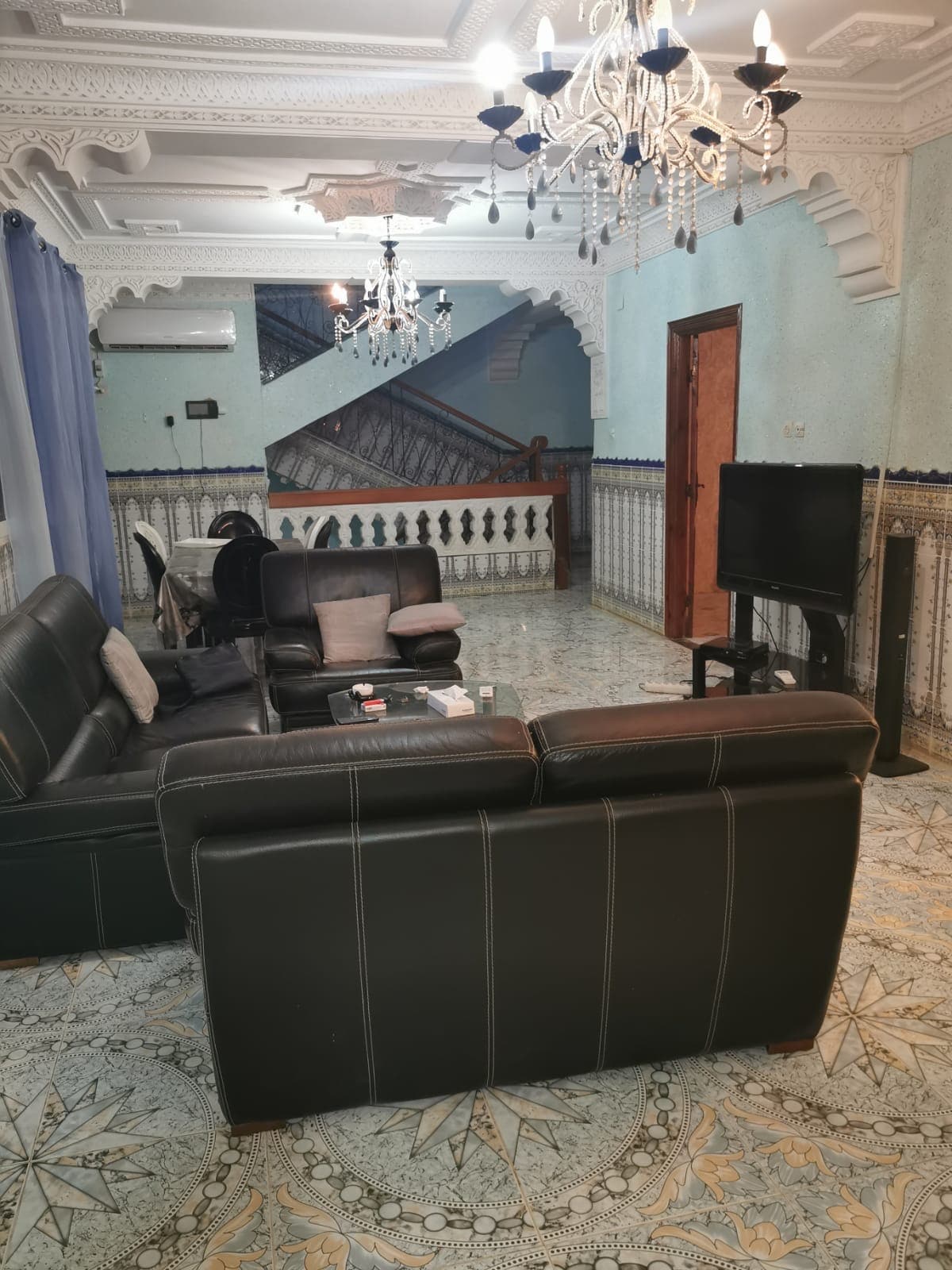 A spacious living area features dark leather seating positioned around a glass table, providing ample space for relaxation. A chandelier hangs from the ornate ceiling, while a television stands on a black cabinet. The tiled flooring adds a touch of elegance to the overall design.