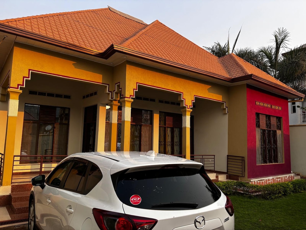 A vibrant exterior of a well-maintained house is showcased, featuring a red and yellow color scheme. The entrance is framed by gracefully designed arches. A white car is parked in the driveway, while green grass surrounds the property.