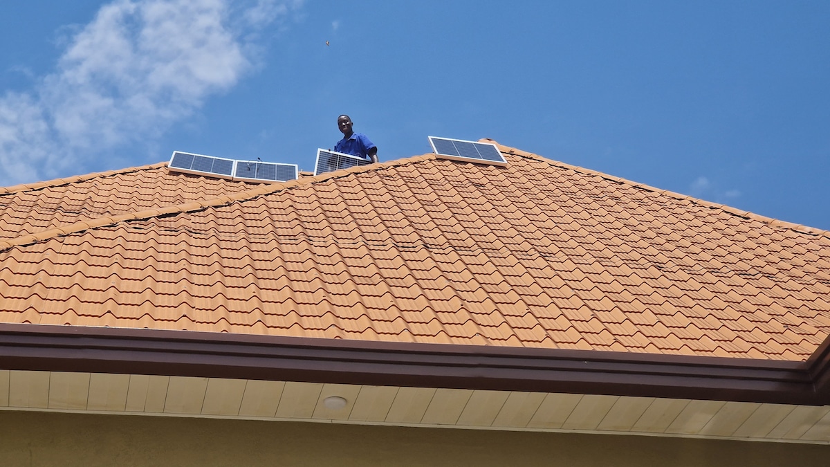 Solar panels are installed on a sloped, tiled roof, contributing to an eco-friendly energy solution. A person is seen working on the roof, which is bathed in sunlight against a clear blue sky.