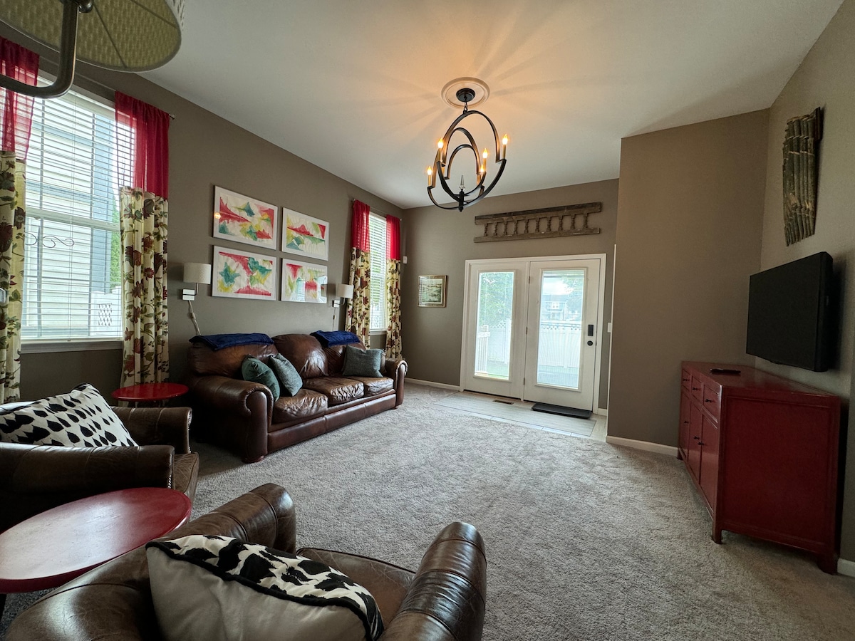 A spacious living area features large windows dressed with colorful curtains, allowing natural light to brighten the space. Comfortable leather sofas and a patterned armchair provide ample seating. A sleek television is mounted on the wall, and a red cabinet adds a pop of color.