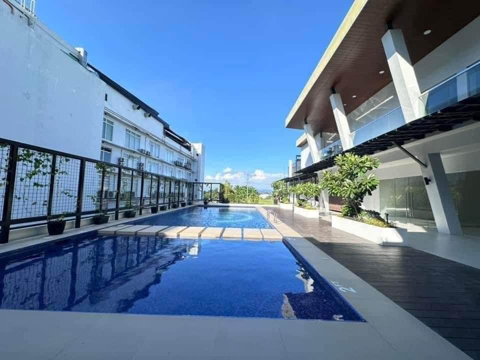 A well-maintained pool area is shown, with a clear blue water reflecting the sky. Surrounding greenery and lounge areas are visible, offering ample space for relaxation. A modern building is situated in the background, enhancing the inviting ambiance of this outdoor space.