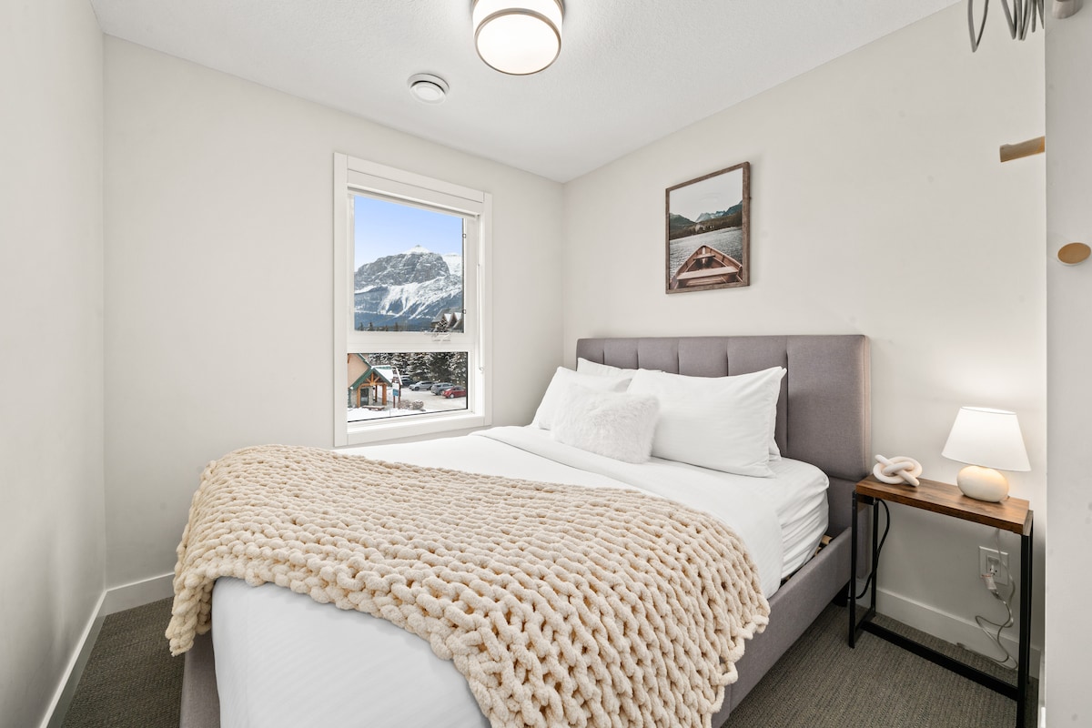 Уютна спалня с голямо двойно легло, изпълнена с естествена светлина, с меки неутрални тонове и прозорец, който показва красив планински пейзаж.