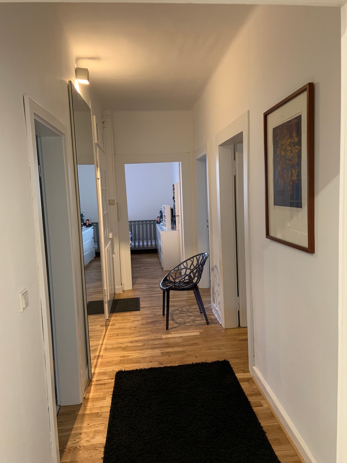 A hallway is shown, featuring light-colored walls and wooden flooring. A large mirror is positioned on one side, enhancing the sense of space. A stylish chair is placed against the wall, with doorways leading to other rooms visible in the background.