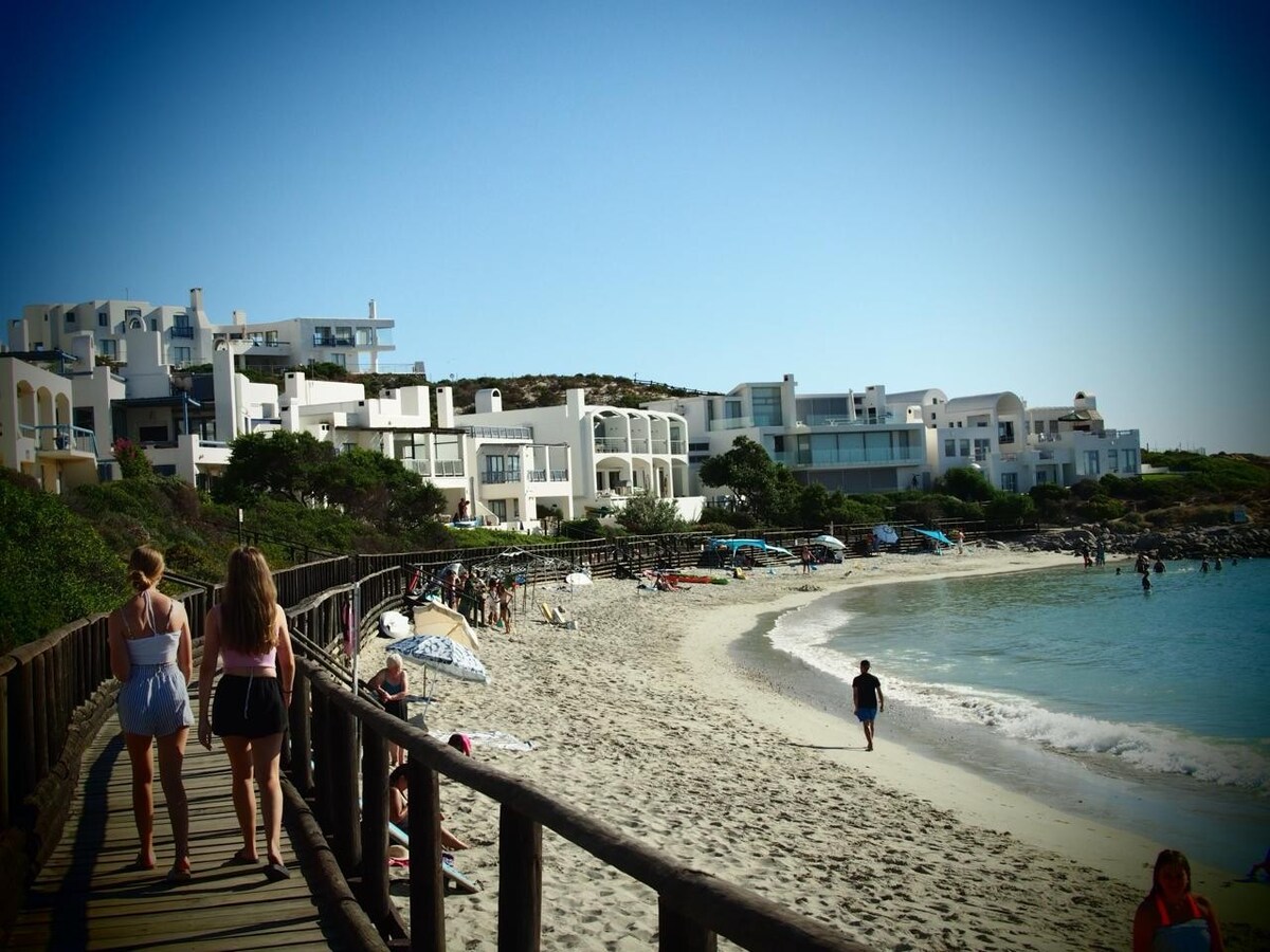 A boardwalk winds along a sandy beach, with visitors walking beside the ocean. The coastline is lined with white buildings, offering a serene coastal backdrop. Sun loungers and umbrellas are set up along the shore, inviting relaxation and enjoyment of the beach environment.