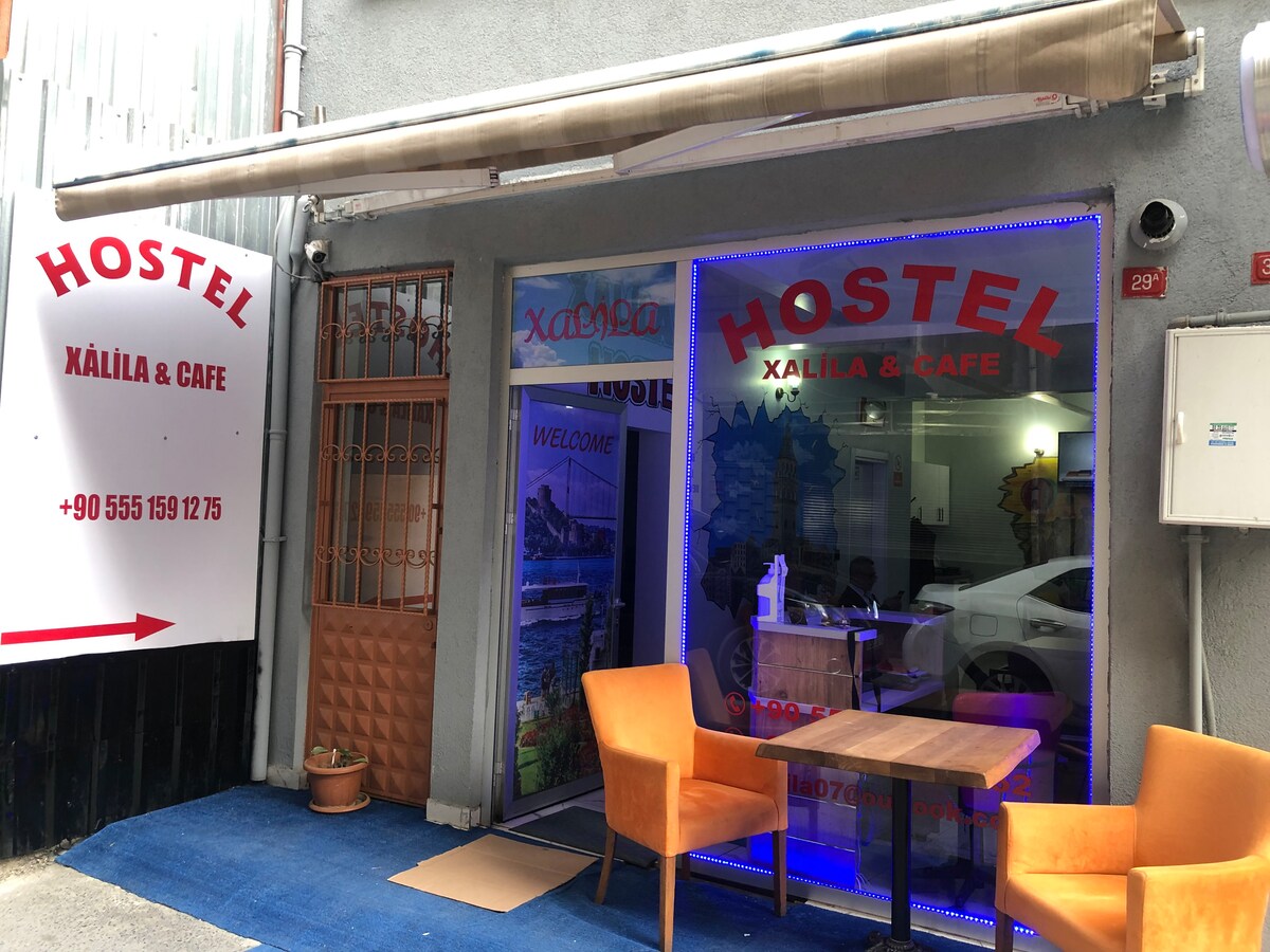 The hostel’s entrance features a welcoming facade with large red lettering spelling 'HOSTEL' and 'WELCOME.' A small outdoor seating area includes two vibrant orange chairs positioned on a blue carpet, inviting guests to relax. A glass door leads into the interior, where amenities continue.