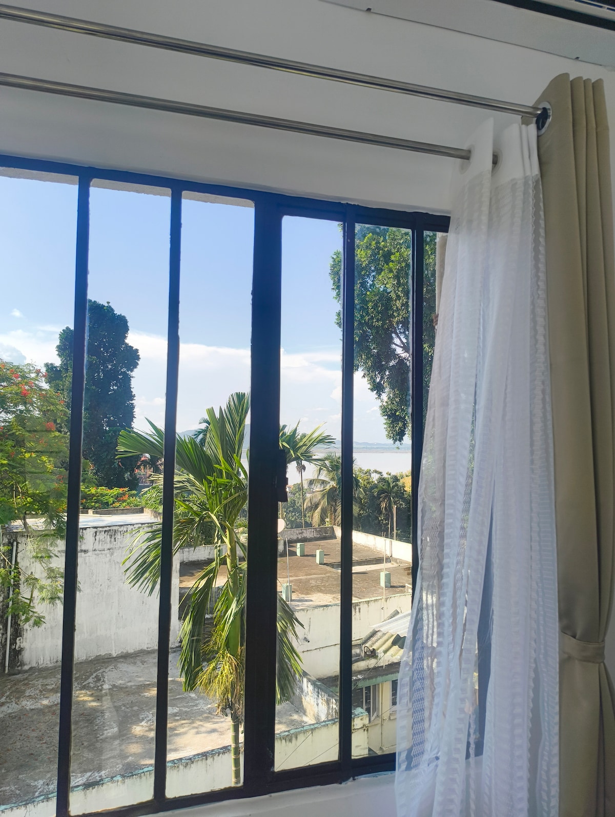 A window with black metal bars offers a view of lush greenery and distant hills. Sunlight filters through sheer white curtains, creating a calm ambiance. The frame is complemented by a glimpse of the sky, showcasing a mix of clouds and blue.