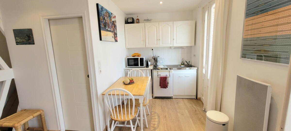A compact kitchen area features white cabinetry and essential appliances, including a microwave, coffee machine, and stove. A small wooden table with white chairs is positioned in front of a window, allowing for natural light. Decorative artwork is displayed on the walls.