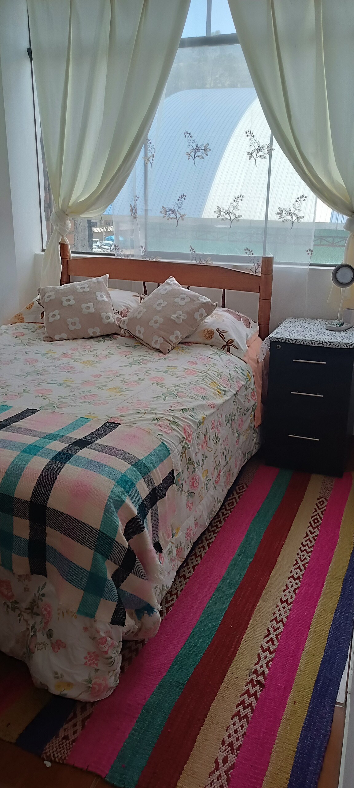 A cozy bedroom features a spacious bed adorned with a flower-patterned blanket and decorative cushions. Natural light filters through sheer curtains, casting a soft glow. A striped rug adds color beneath the bed, complementing a bedside table with a lamp.