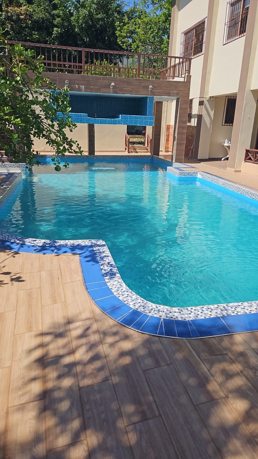 A refreshing swimming pool is surrounded by a tiled deck, featuring a vibrant blue edge. Sunlight reflects off the water, creating a serene environment. In the background, a balcony overlooks the pool area, providing additional seating options.