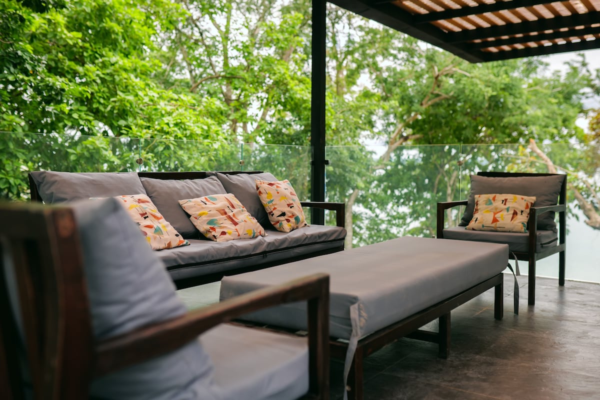 A covered terrace is furnished with a spacious couch and two matching chairs, all featuring colorful cushions. Large glass panels provide unobstructed views of the surrounding greenery, creating a serene outdoor space for relaxation.