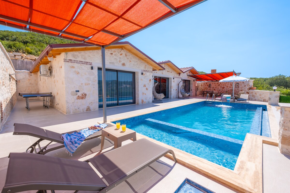 A refreshing swimming pool features crystal-clear water, bordered by a spacious terrace. Chaise lounges are positioned nearby, providing a relaxing spot under shaded awnings. The stone exterior of the house complements the outdoor setting, with large glass doors offering views into the inviting interior.
