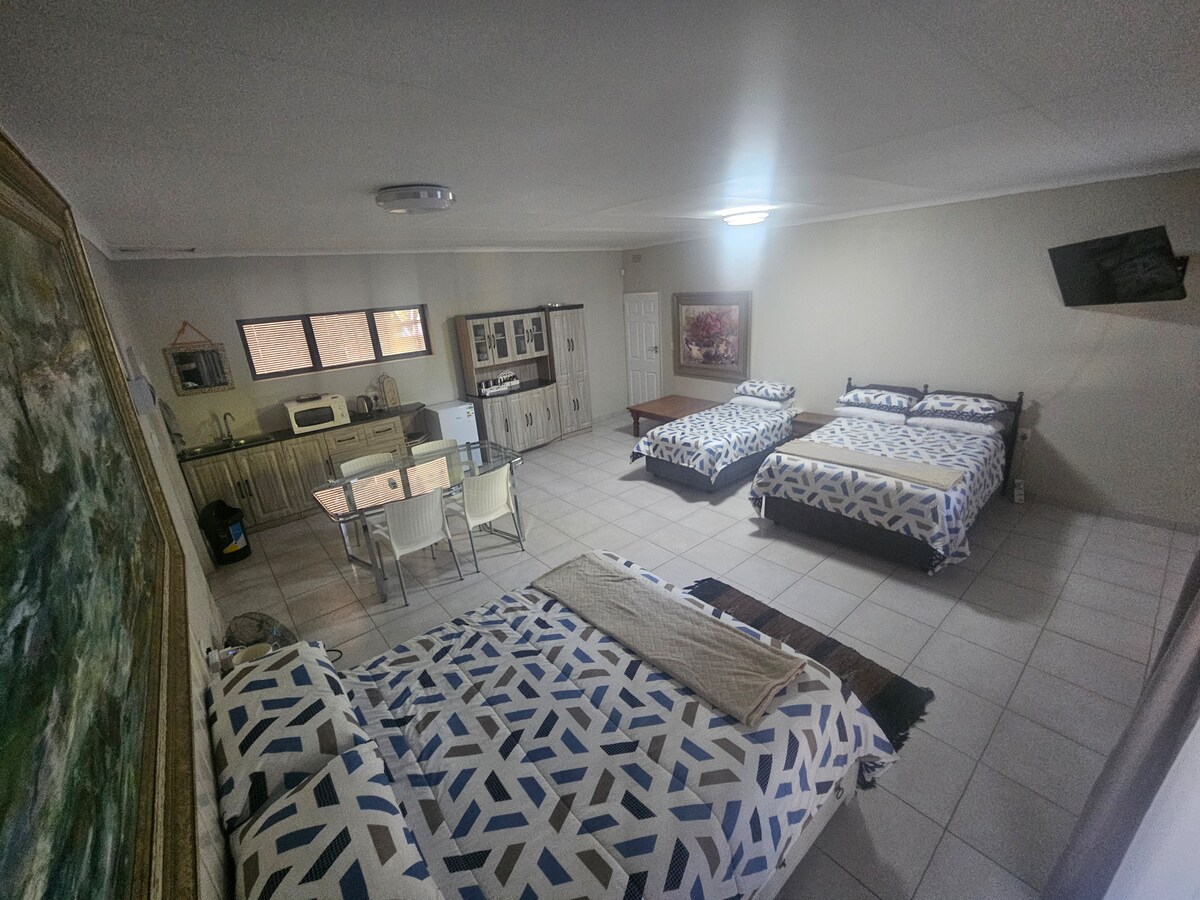 A spacious room is illustrated with two beds draped in patterned bedding. A kitchenette with cabinetry and a microwave is visible, along with a dining table and chairs. A television is mounted on the wall, and natural light filters through windows, illuminating the area.