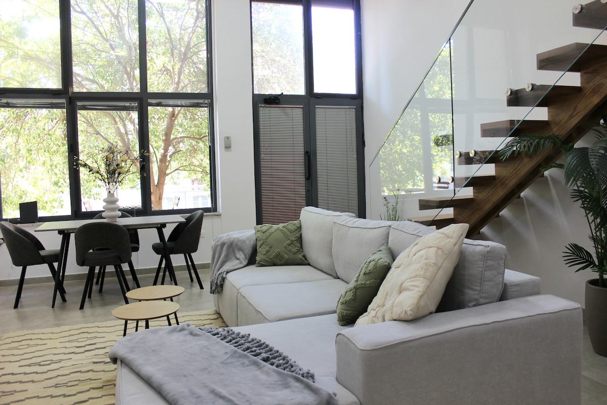 A spacious living area features a large sectional sofa adorned with cushions and a throw blanket. A dining table with chairs is visible, alongside a staircase with wooden steps and glass railings. Natural light streams through tall windows overlooking greenery.
