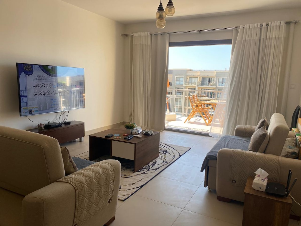 A bright living area features a large TV mounted on the wall and is furnished with two sofas, one of which is a sofa bed. Natural light floods the space through glass doors, leading to a balcony with outdoor seating.