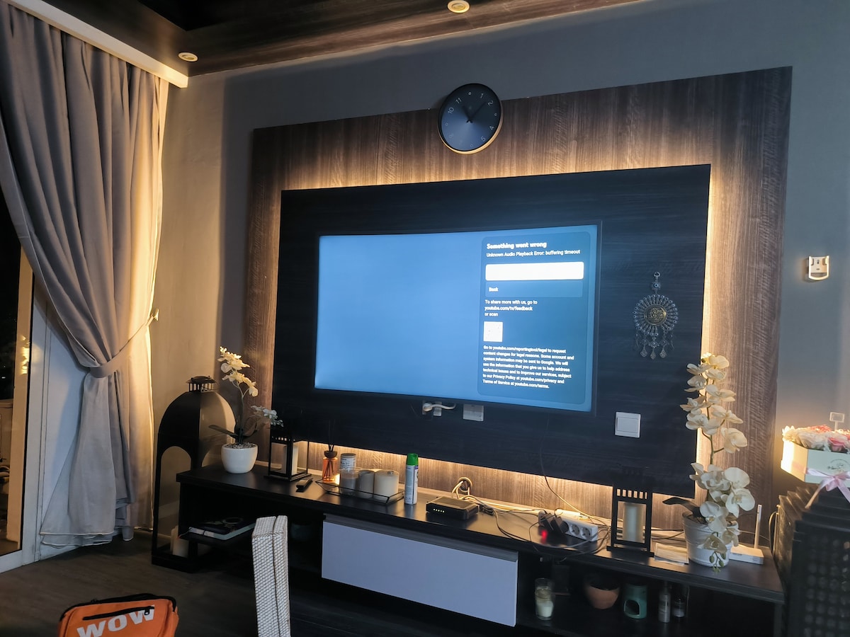 A modern entertainment center is displayed, featuring a large flat-screen television mounted on a dark wooden wall. Decorative plants and elegant vases are arranged on the console below, while soft lighting creates a welcoming ambiance. A round wall clock is positioned above the television.