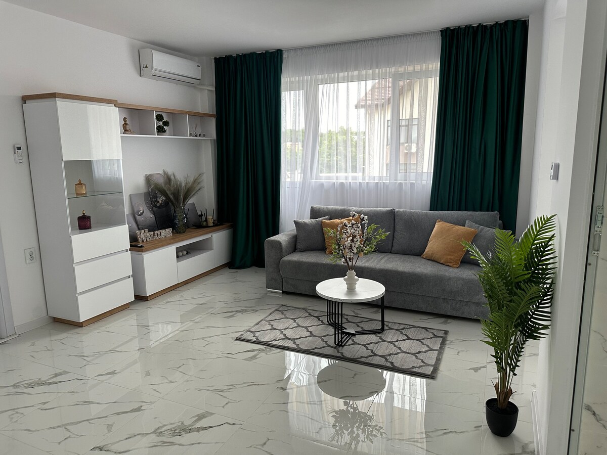 A modern living area showcases a gray sectional sofa adorned with decorative pillows. A round coffee table sits on a geometric rug, framed by large windows draped in sheer curtains. A plant and decorative items are placed on the wall unit, enhancing the space's aesthetic.
