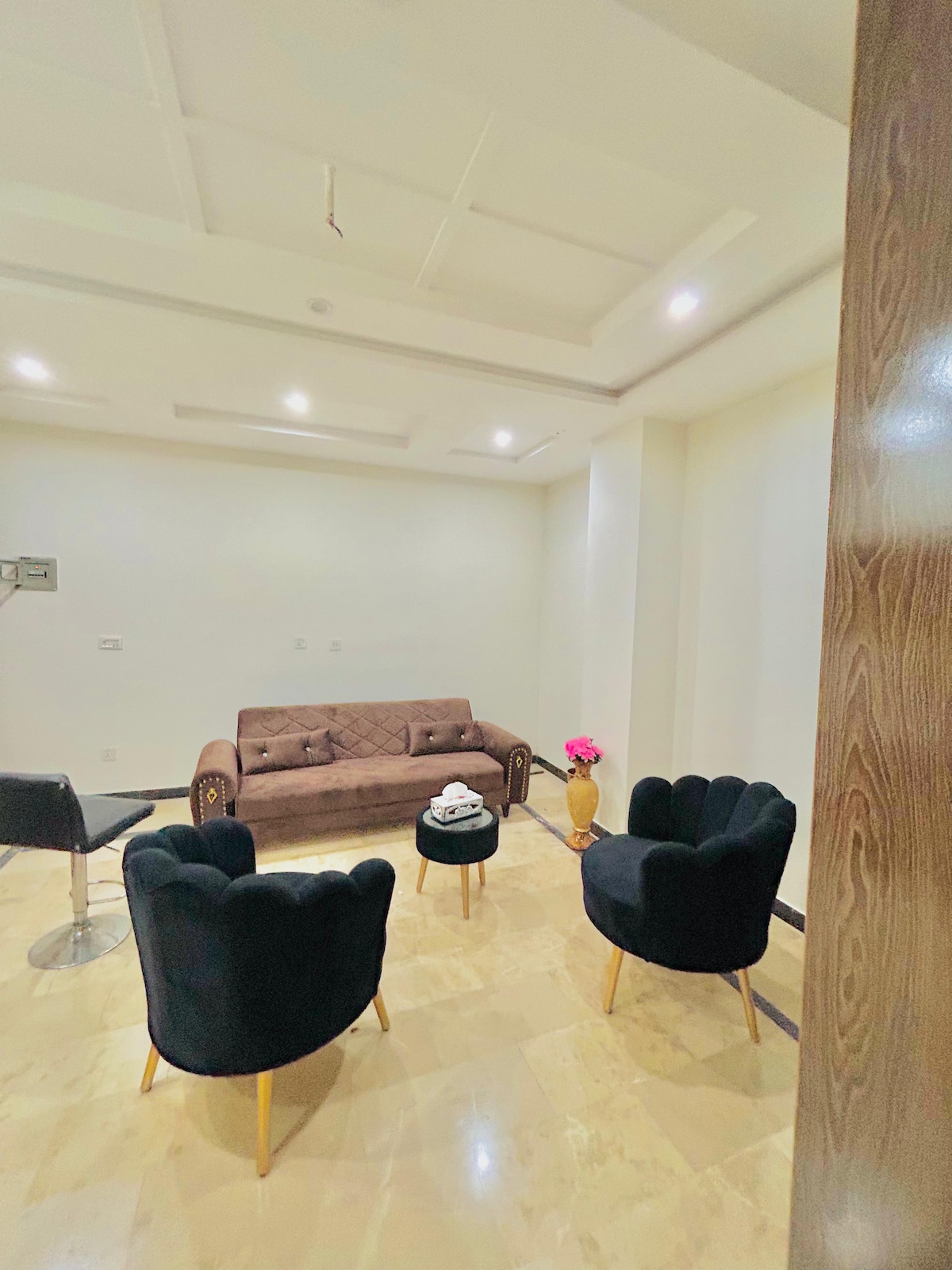 A clean and modern living area features a brown sofa with a quilted design, accompanied by two black velvet chairs with wooden legs. Soft lighting is provided by recessed ceiling lights, creating a calm atmosphere. A small side table holds a decorative vase with flowers.