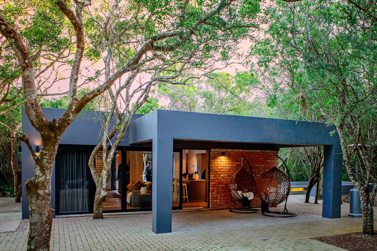 The exterior of a modern structure is framed by lush greenery, with large glass doors allowing visibility into the interior. Two unique hanging chairs are positioned on the patio, offering a comfortable spot to enjoy the natural surroundings.