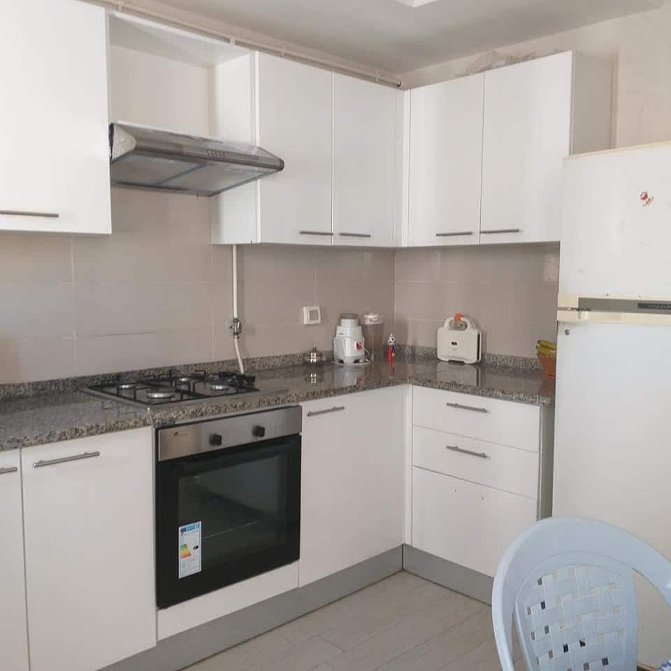 A modern kitchen is equipped with white cabinetry and a granite countertop. A gas stove and an oven are integrated into the kitchen layout. A refrigerator stands nearby, while a small appliance station is placed on the countertop, contributing to a functional cooking space.