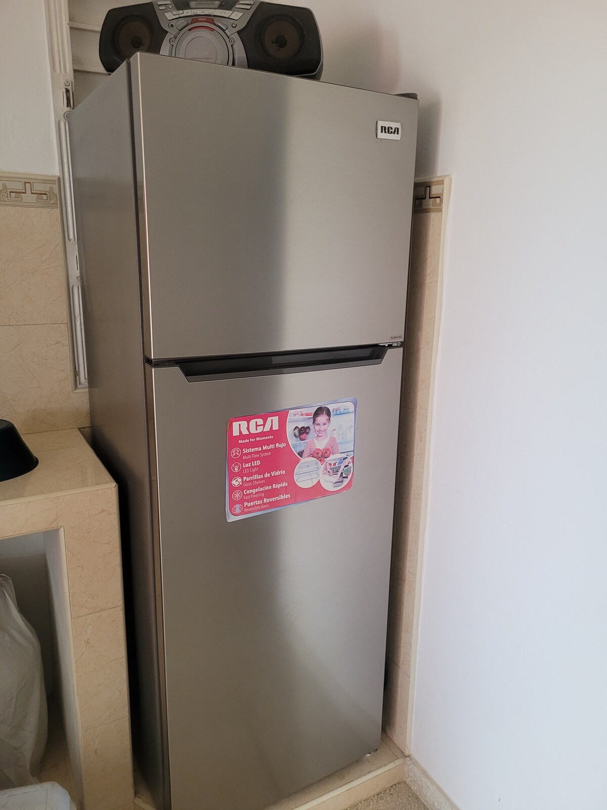A modern stainless steel refrigerator stands against a light-colored wall, featuring a prominent RCA logo on the front. The kitchen area is subtly illuminated, with a countertop visible beside the refrigerator. A compact stereo system is placed above the refrigerator, enhancing the space.
