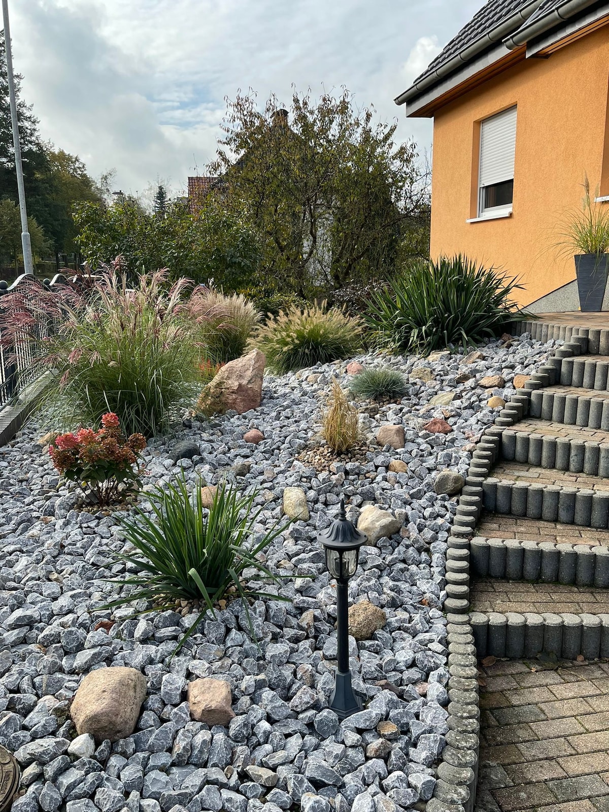 A landscaped garden features an array of decorative stones and plants, creating a natural aesthetic. The pathway, lined with pavers, leads to the entrance of the building. Various shrubs and ornamental grasses add texture, complemented by a subtle lighting fixture positioned nearby.