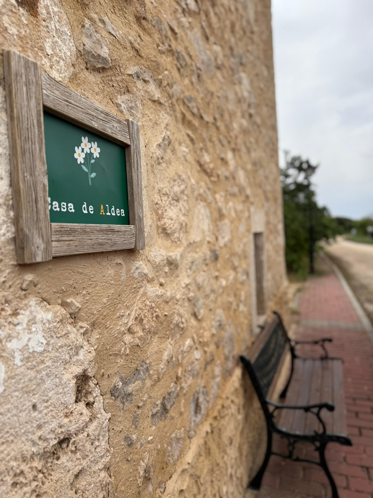 The exterior of Casa de Aldea is shown, featuring a rustic stone wall and a wooden sign with floral design. A bench with a traditional design is positioned nearby, providing a space for relaxation. Natural surroundings complement the tranquil setting.