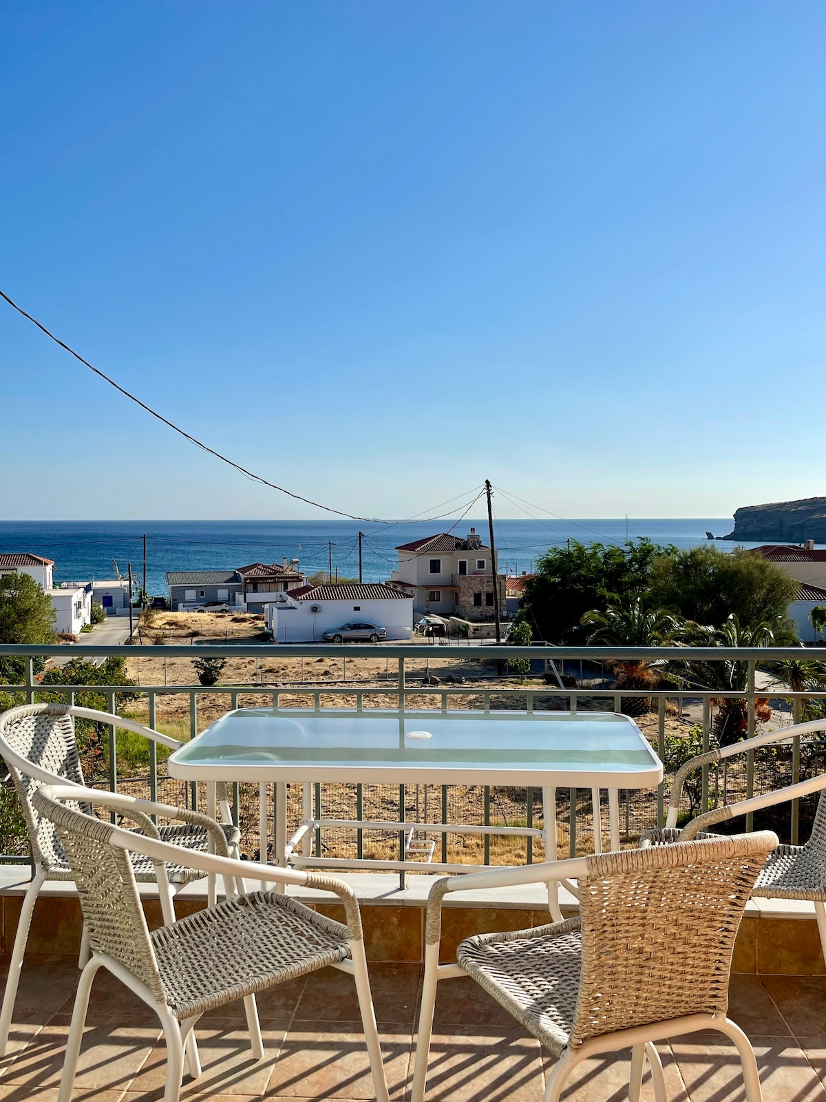 A private balcony features a table and four chairs, overlooking a serene view of the Aegean Sea. The landscape includes distant houses and green foliage, set against a clear blue sky, creating a tranquil spot for relaxation.