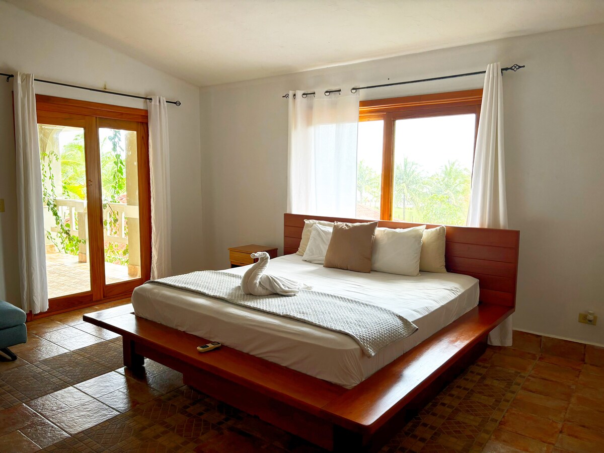 A bright sleeping area features a spacious bed with neutral-colored bedding and decorative pillows. Large windows allow ample natural light, complemented by sheer white curtains. A wooden accent wall adds warmth, while a small nightstand sits beside the bed.
