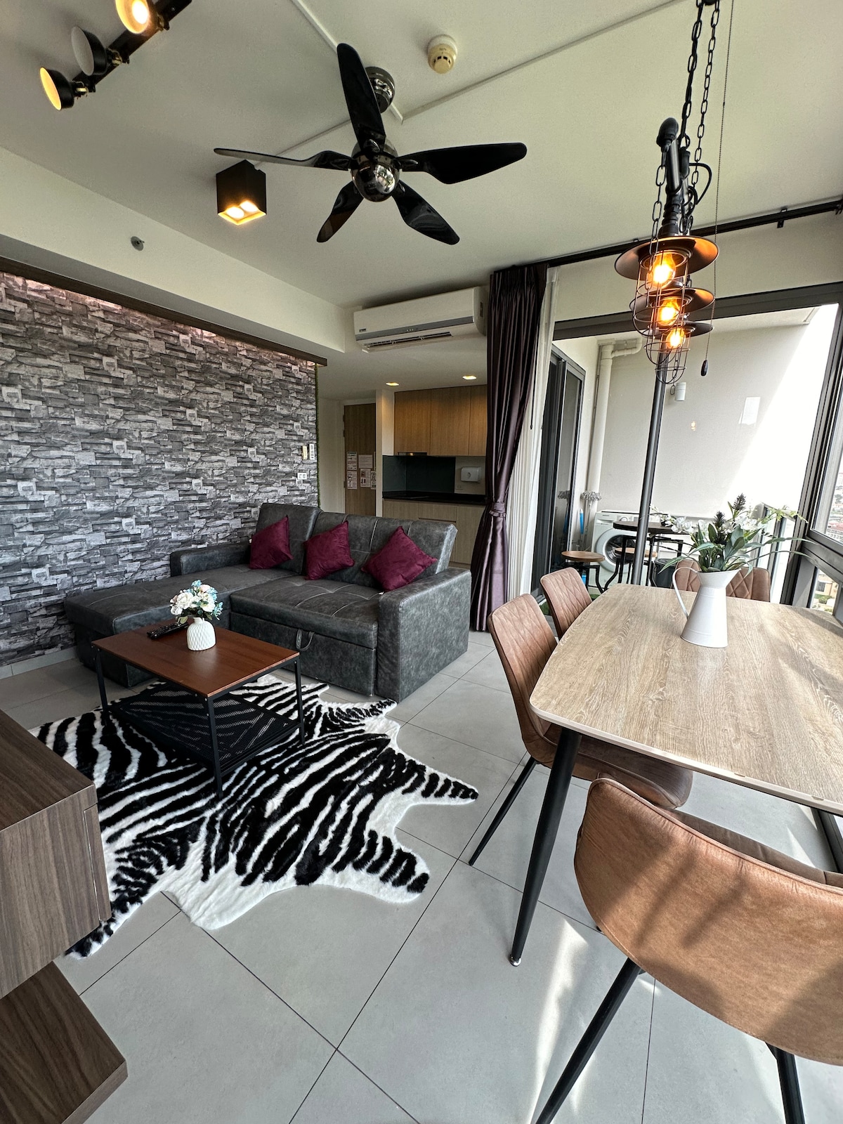 A modern living space is highlighted by a textured stone accent wall. A gray sofa with burgundy cushions faces a wooden coffee table. A dining area with stylish chairs sits nearby, surrounded by large windows that allow ample natural light.