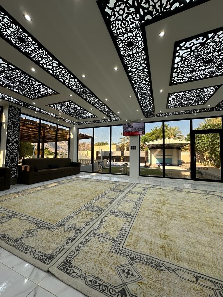 A spacious lounge area features intricate ceiling designs and large windows that provide natural light and views of the outdoor greenery. Plush seating is arranged on a beautifully patterned rug, enhancing the elegance of the room.