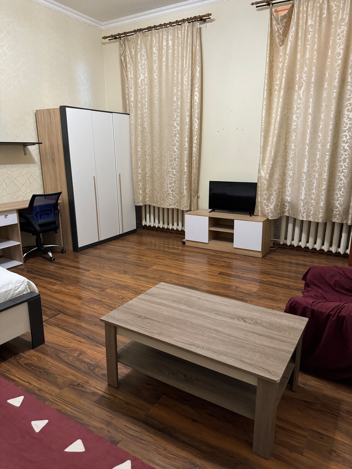 A spacious room featuring warm wooden flooring and light-colored walls. A small table is positioned centrally, surrounded by seating. A wardrobe stands against one wall, while a TV unit and a television are located nearby. Natural light filters through curtains.