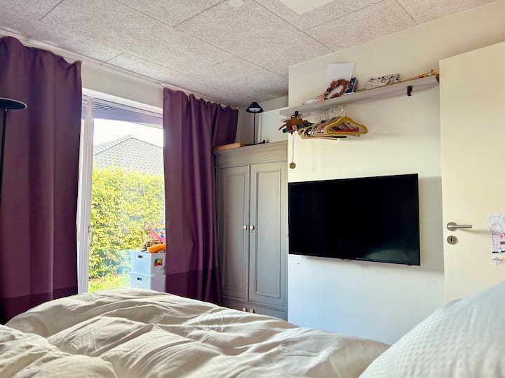 חדר שינה מואר בגודל 10.5 מ"ר עם מיטה זוגית, טלוויזיה תלוית קיר, Chromecast וארון בגדים. וילונות האפלה ונוף לגינה כדי להירגע באופן אופטימלי.