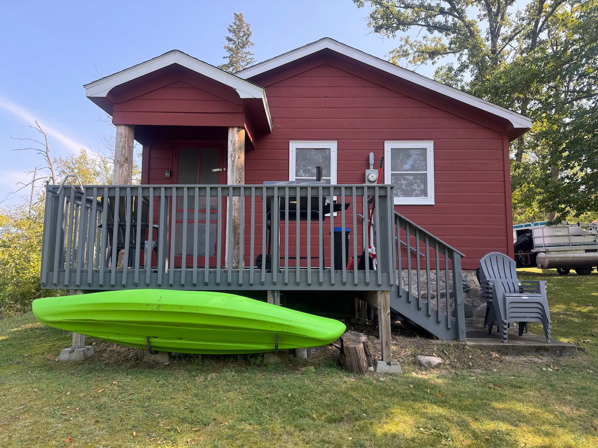A rustic red cabin is depicted with a wooden deck and steps leading to the entrance. A bright green kayak rests against the lower side of the cabin. Two outdoor chairs are positioned on the deck, surrounded by grass and trees in the background.