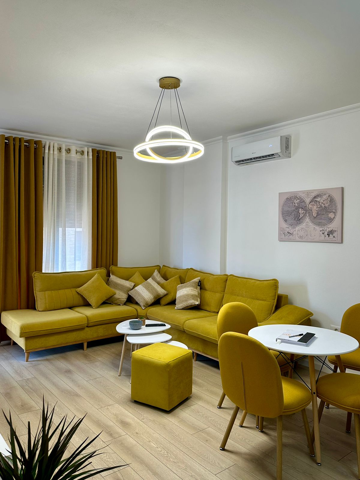 A modern living area is highlighted by a sectional sofa in a warm yellow hue, adorned with decorative cushions. A round white table and chairs complement the space, with a stylish ceiling light providing illumination. Soft natural light filters through the sheer curtains.