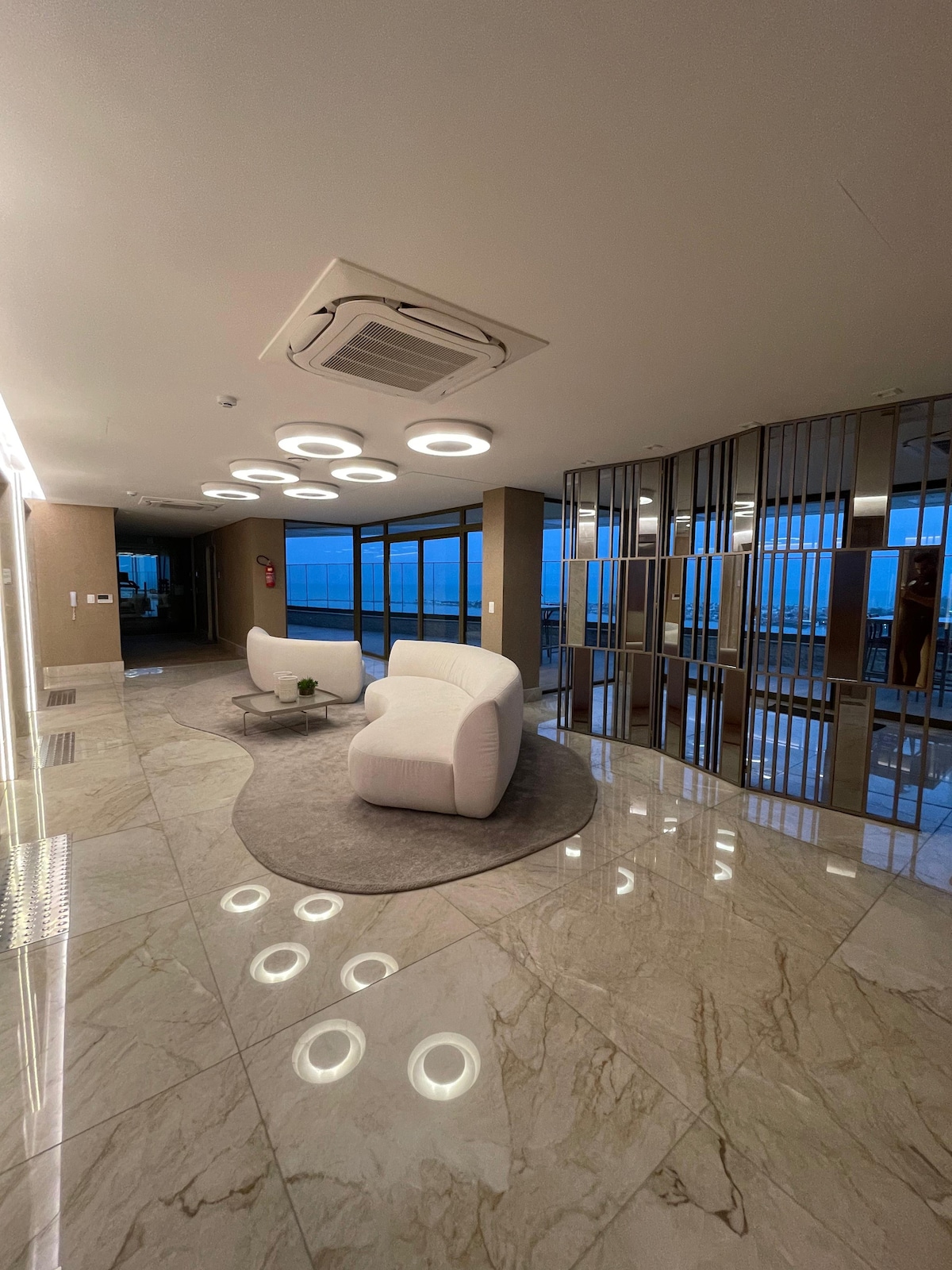 A modern lobby area is presented with glossy marble floors and contemporary furnishings. A pair of white armchairs is situated on a circular gray rug. Large glass windows allow natural light to fill the space, providing expansive views of the surroundings.