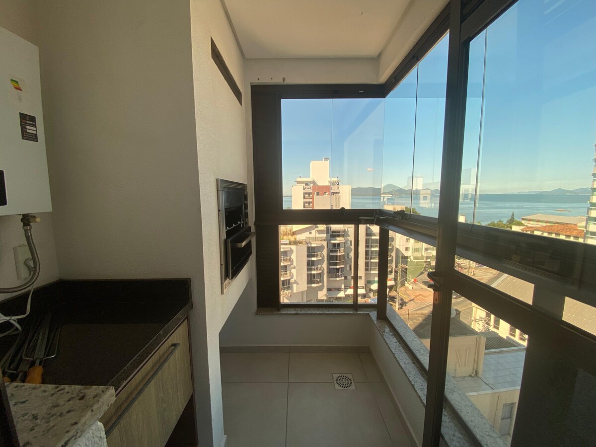 A well-appointed kitchenette is showcased, featuring modern appliances and a granite countertop. Large windows provide a view of the surrounding area and the distant water. Natural light fills the space, enhancing the clean lines and functional layout.