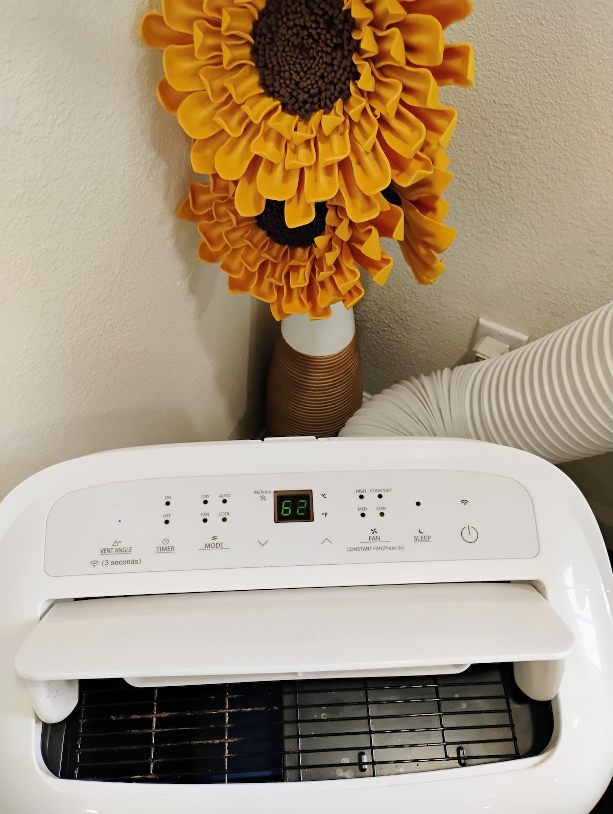 An air fryer is displayed prominently, featuring a digital control panel with various settings. The sleek white appliance sits beneath a decorative sunflower plant that adds a touch of color. A gray ventilation hose is visible in the background, contributing to the kitchen's functionality.