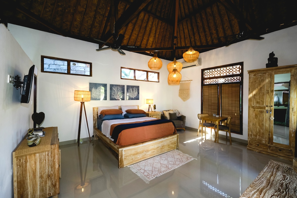 The bedroom features a spacious bed with a warm color palette and natural wooden furnishings. Large windows allow for natural light, while unique pendant lighting enhances the ambiance. A wooden wardrobe and a small table are visible, along with polished flooring that complements the tropical theme.