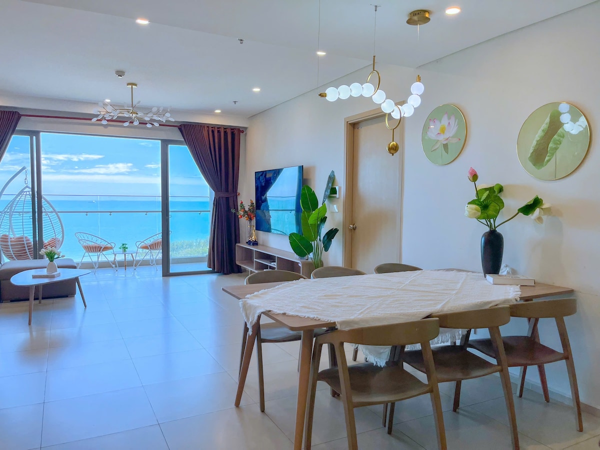 A bright living area is shown, featuring a dining table with six chairs and a television on a media console. Large glass doors open to a balcony with sea views, while potted plants and decorative wall art add a touch of greenery and elegance.