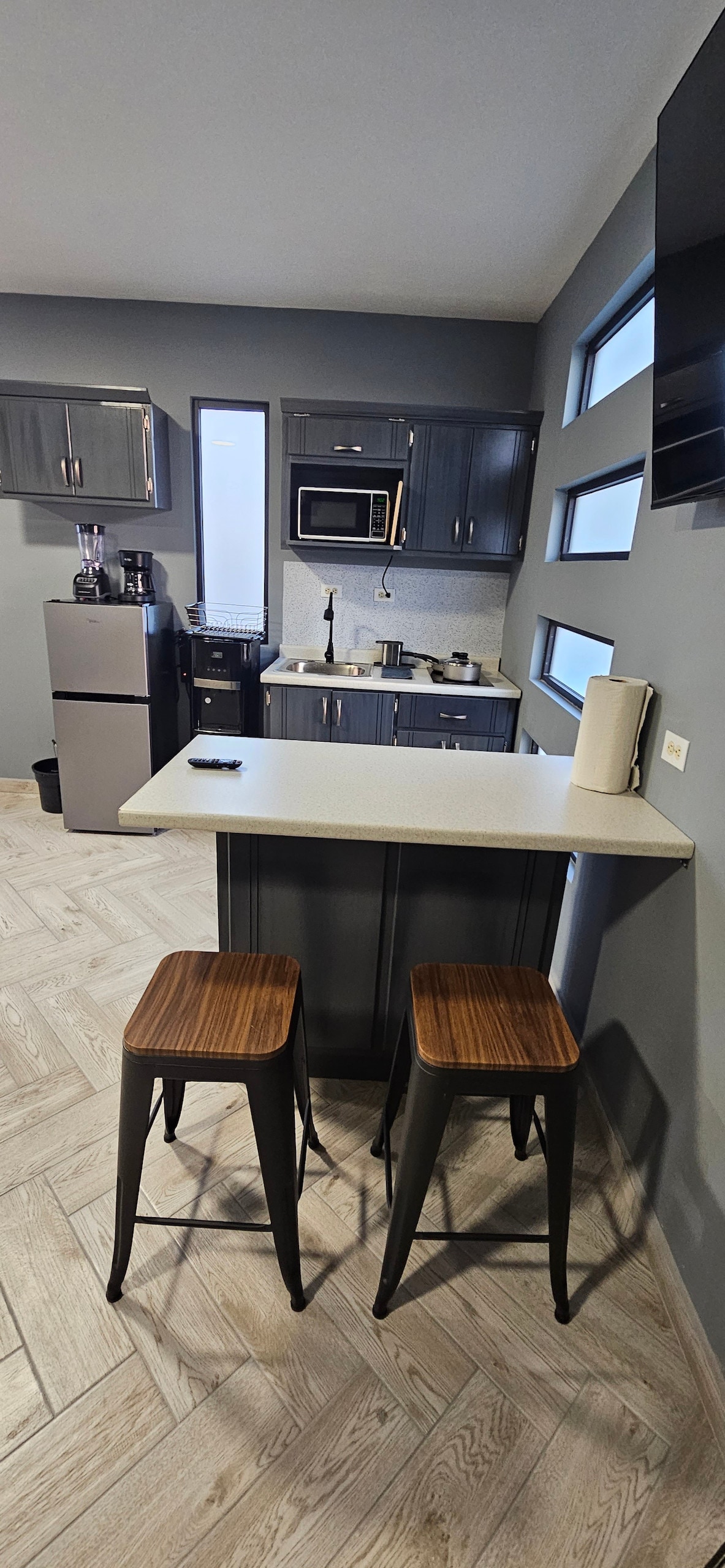 A compact kitchen area is equipped with modern appliances, including a mini refrigerator and microwave. Two metal stools sit at a small, table-like counter. Natural light enters through multiple windows, highlighting the contemporary cabinetry and the textured floor design.