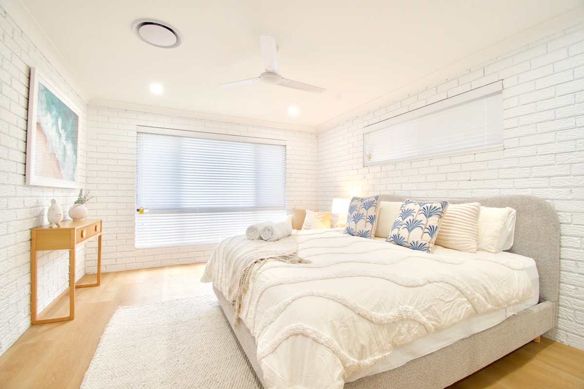 A spacious bedroom with light wood flooring and white brick walls creates a calming environment. A large bed is dressed with soft linens and decorative pillows. Natural light floods the room through large windows with blinds, and a small wooden console table complements the decor.