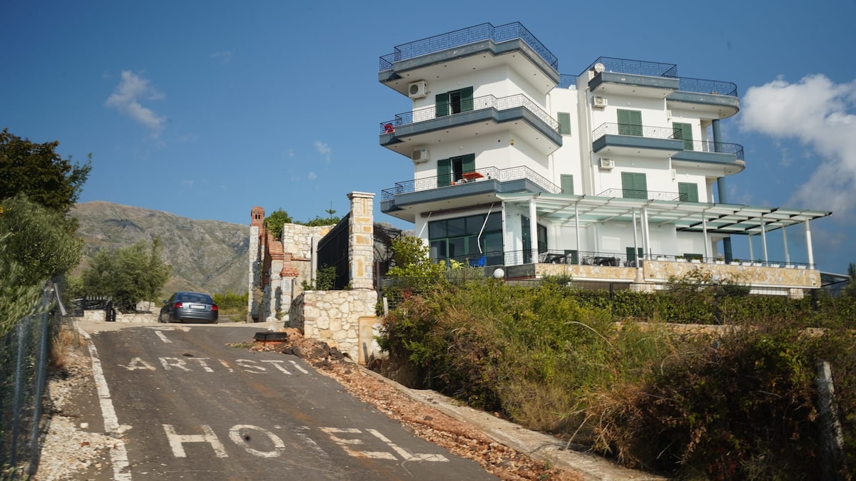 The hotel, featuring a modern design, is positioned on a hillside with panoramic views. A spacious terrace extends from the glass-enclosed common area. A private parking lot is visible, alongside a dirt road leading to the entrance. Lush greenery surrounds the property.