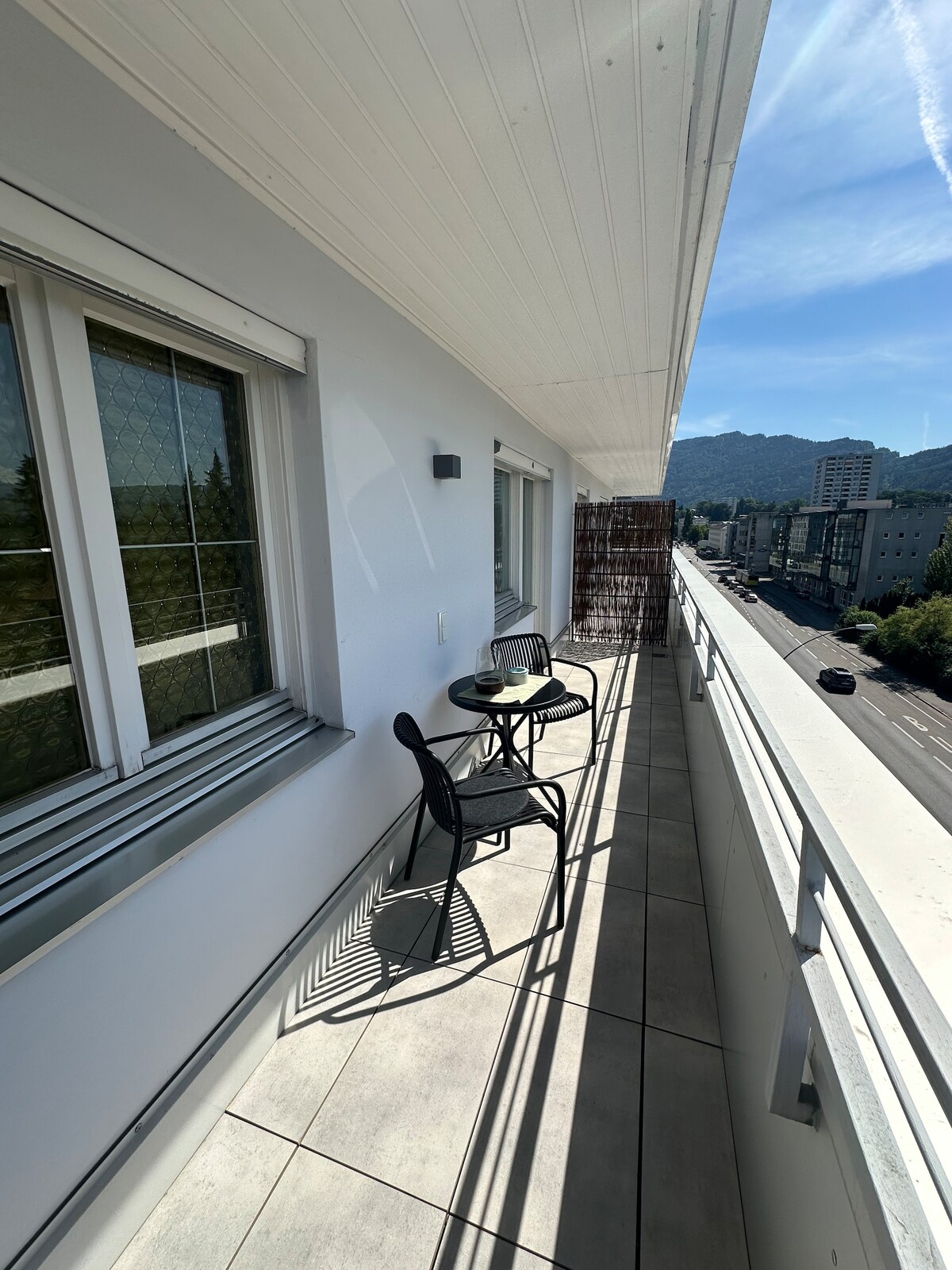 A balcony area features a small round table and two chairs, offering a comfortable place to enjoy outdoor views. Sunlight creates distinct shadows on the tiled floor, with a clear view of the surrounding buildings and mountains in the background.