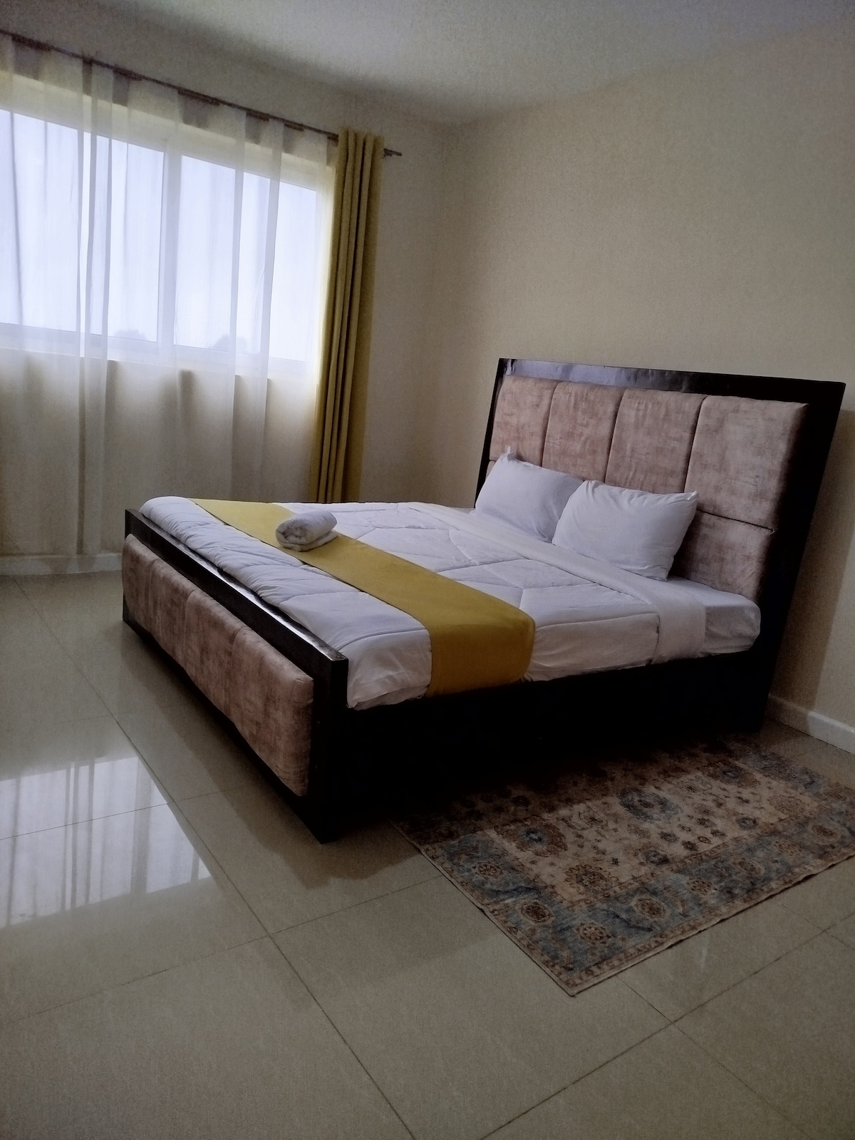 A simple bedroom features a spacious bed with a soft headboard, draped in crisp white linens and accented by a yellow runner. Natural light filters through sheer curtains, illuminating the glossy tiled floor and a small area rug underneath the bed.