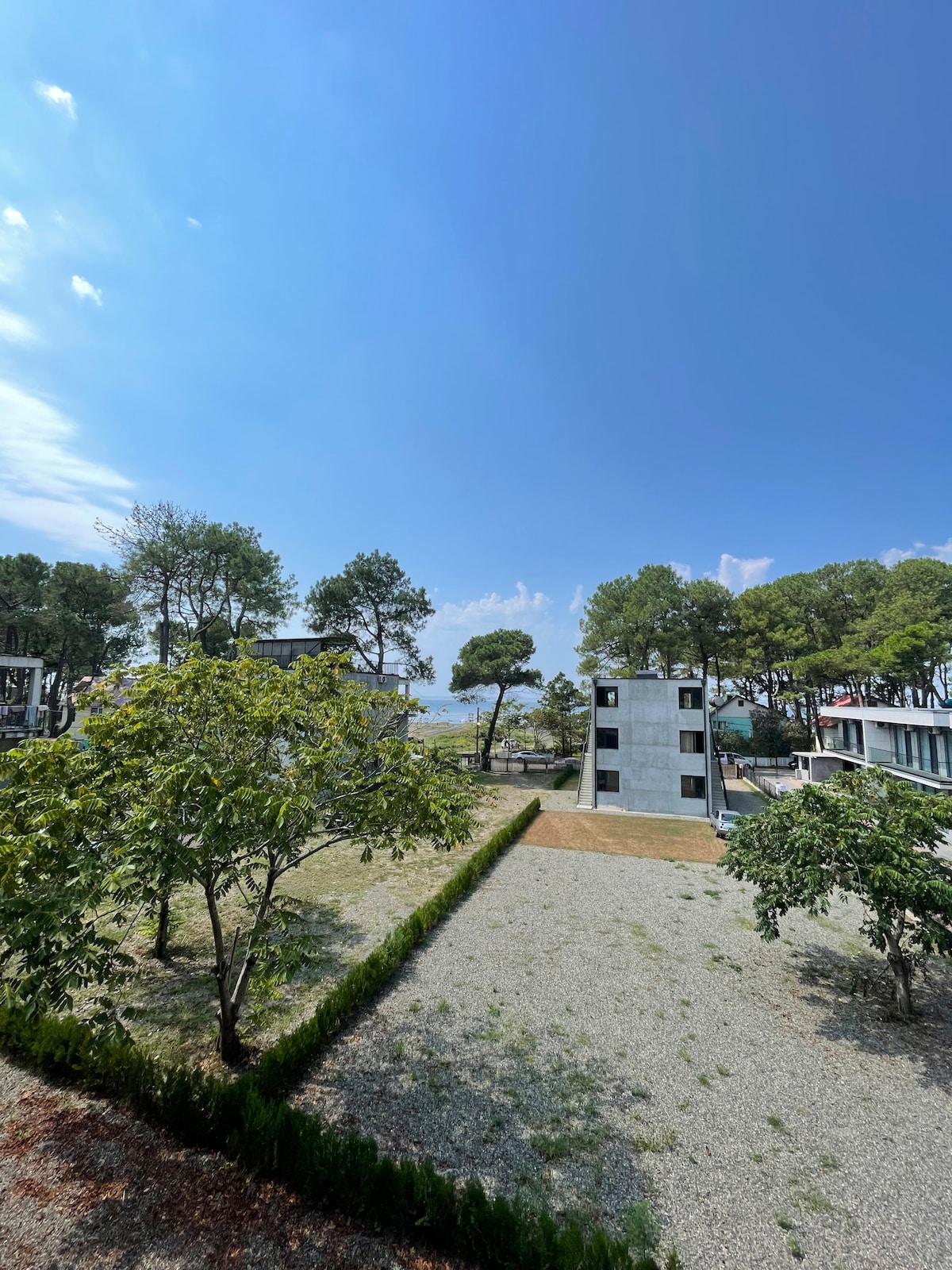 An open landscape features a mix of grassy areas and pebble pathways, bordered by various trees. A modern building stands in the background, framed by blue skies and distant hills, creating a serene environment for family gatherings.