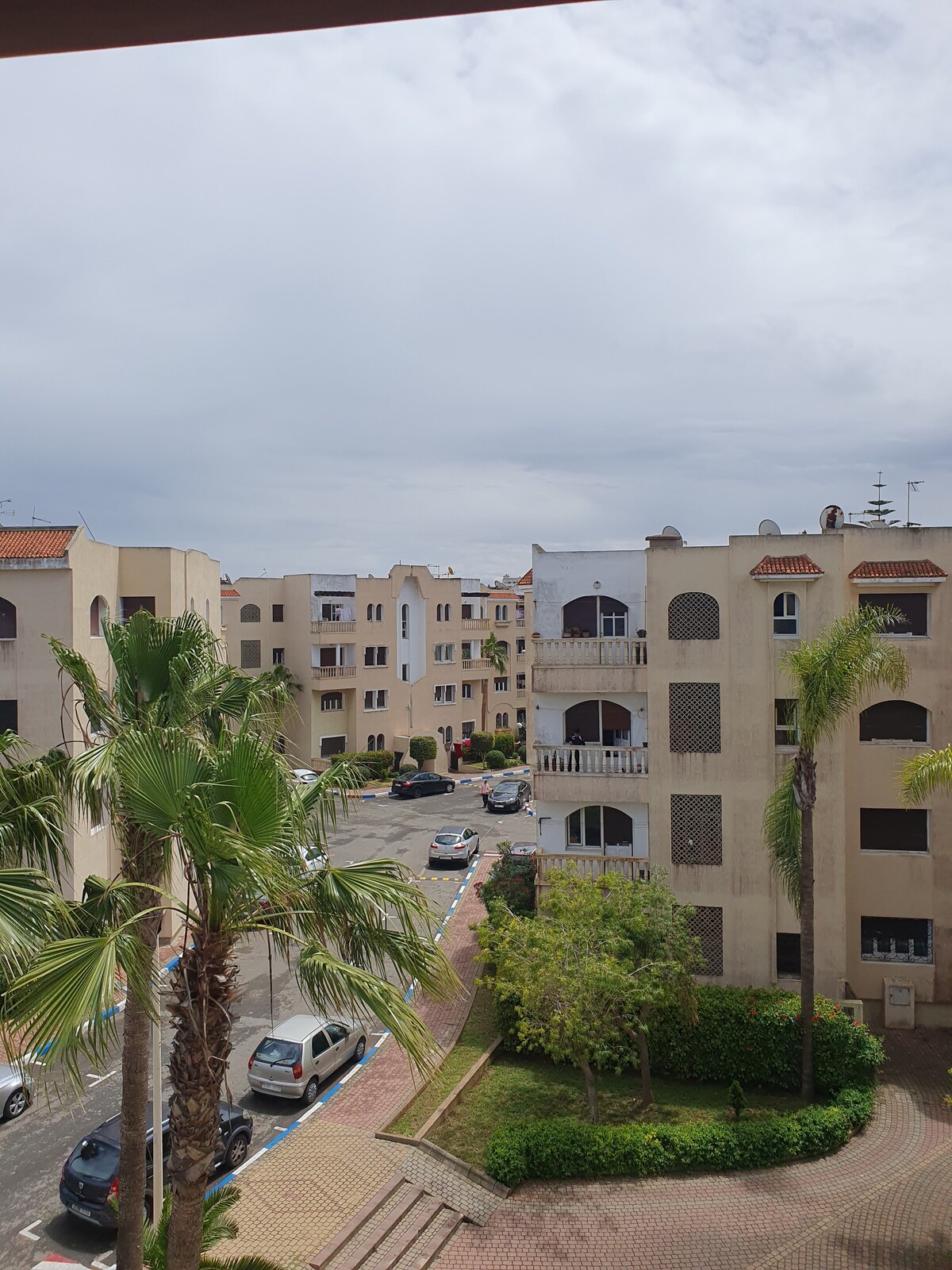 A view from the balcony captures a residential area with multiple buildings in soft beige tones. Lush palm trees and greenery are visible in the foreground, while parked cars are lined along the driveways. The sky is partly cloudy, adding a serene backdrop to the scene.